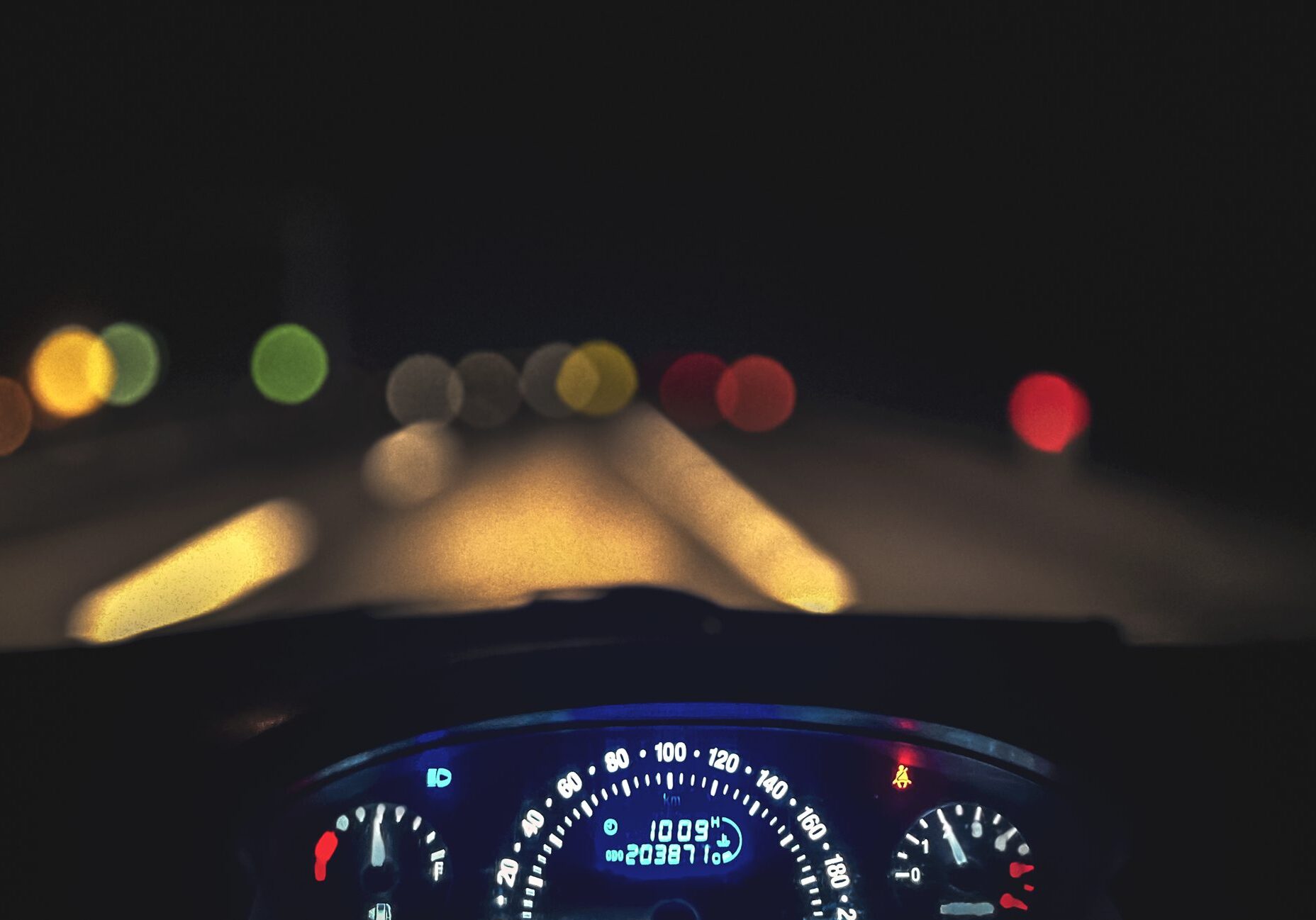 Blurred night view from a car dashboard showing illuminated speedometer and road lights.